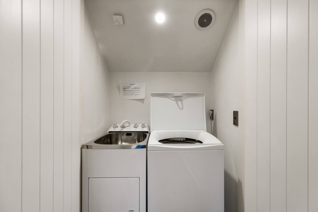 A laundry room with a front-loading washer and dryer, a sink, and a ventilation system.