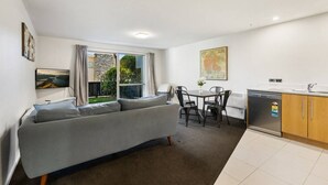 A compact living space with a sofa, dining table, chairs, a kitchen area with a dishwasher, and a large window with curtains. - Lake & Alps on Your Doorstep | Resort Facilities (Wānaka)