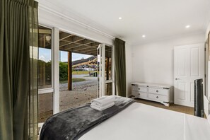 Un dormitorio con cama, cómoda, televisor y puerta corrediza de vidrio que da a vista de una carretera y edificios.