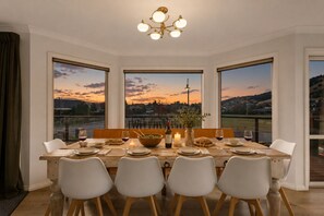 A dining room with a large table set for six, large windows with a view of the countryside, and a chandelier. - Timberstone House | Spacious 5BR, Walk to Town (Wānaka)