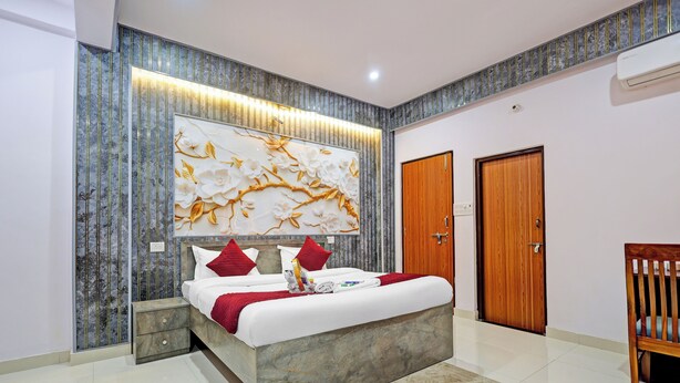 A modern bedroom with a large bed, a decorative wall panel, a wooden door, and a chair. - Hotel Rahi Residency (Varanasi)