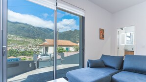 A modern living room with a large sliding glass door leading to a balcony, a blue sofa, and a view of a mountainous landscape. - Casa Barranca by Atlantic Holiday (São Vicente)