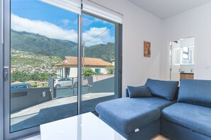 Ein modernes Wohnzimmer mit einem großen Schiebeglasfenster, das auf einen Balkon führt, einem blauen Sofa und Blick auf eine Berglandschaft.