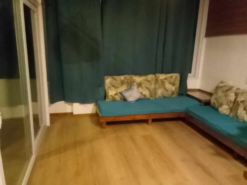 Una habitación con piso de madera, un sofá con cojines estampados y una ventana con cortinas.