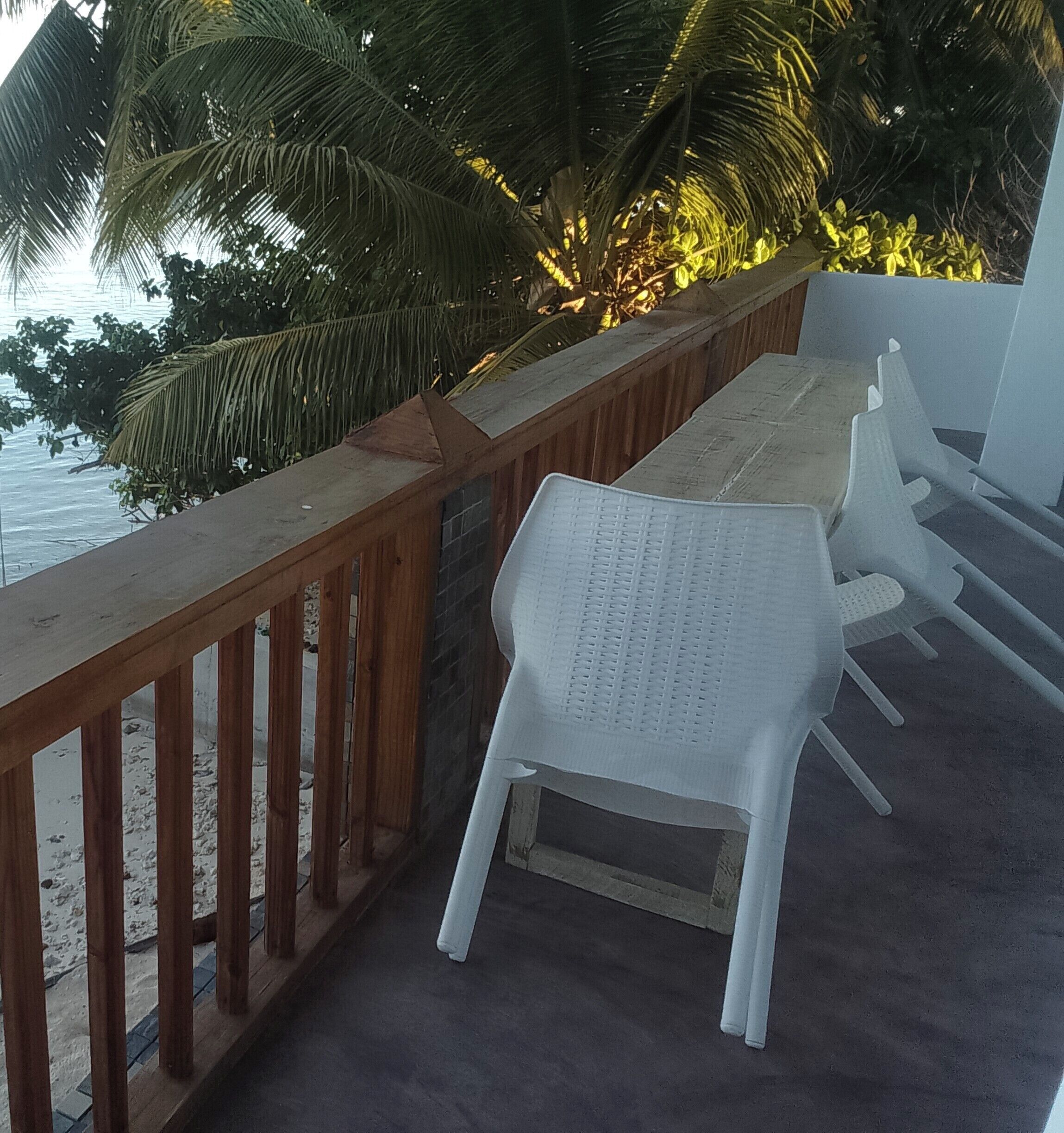 A balcony with a wooden railing, white plastic chairs, and a view of the sea and palm trees.