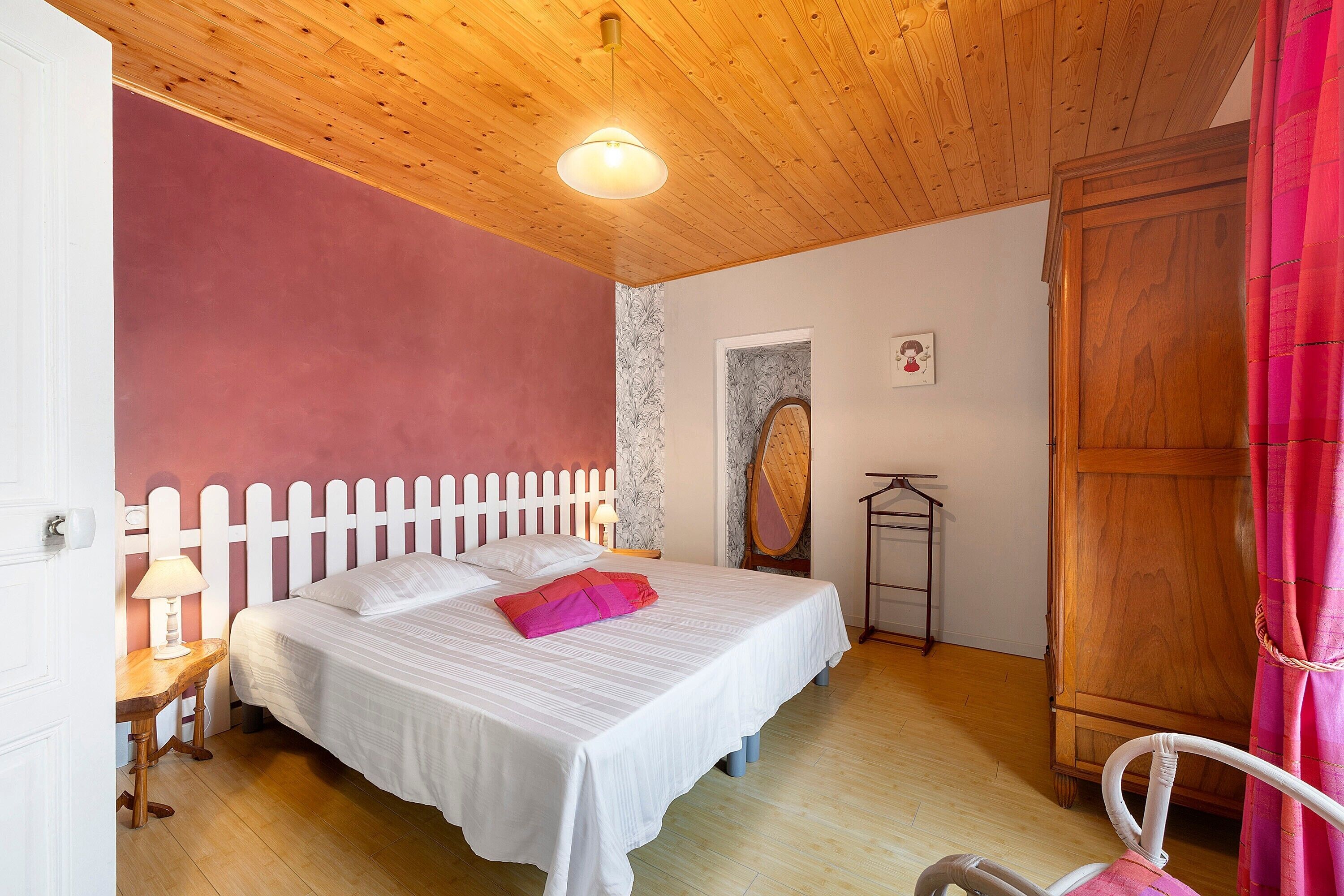 A bedroom with a wooden ceiling, a white bed with pillows, a nightstand, a wooden wardrobe, and a door with a white picket fence.