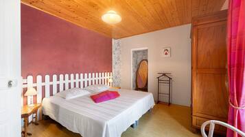 A bedroom with a wooden ceiling, a white bed with pillows, a nightstand, a wooden wardrobe, and a door with a white picket fence.