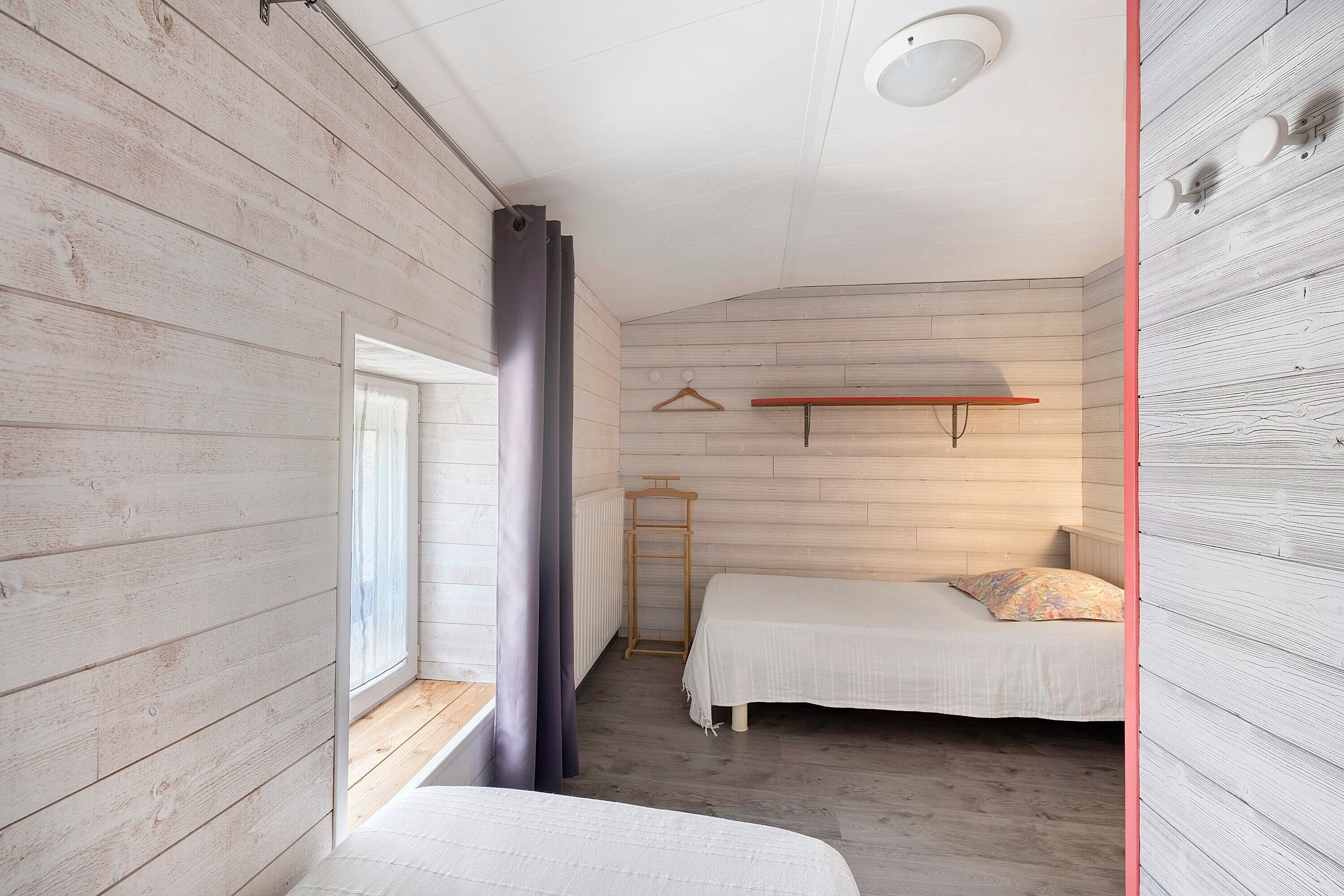 A room with wooden walls, a bed, a window with curtains, a radiator, and a shelf with hangers.