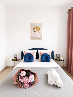 Et pænt opredt soveværelse med seng, puder, et natbord, en lampe, et indrammet kunstværk, en blomsterbuket og sammenrullede håndklæder.
