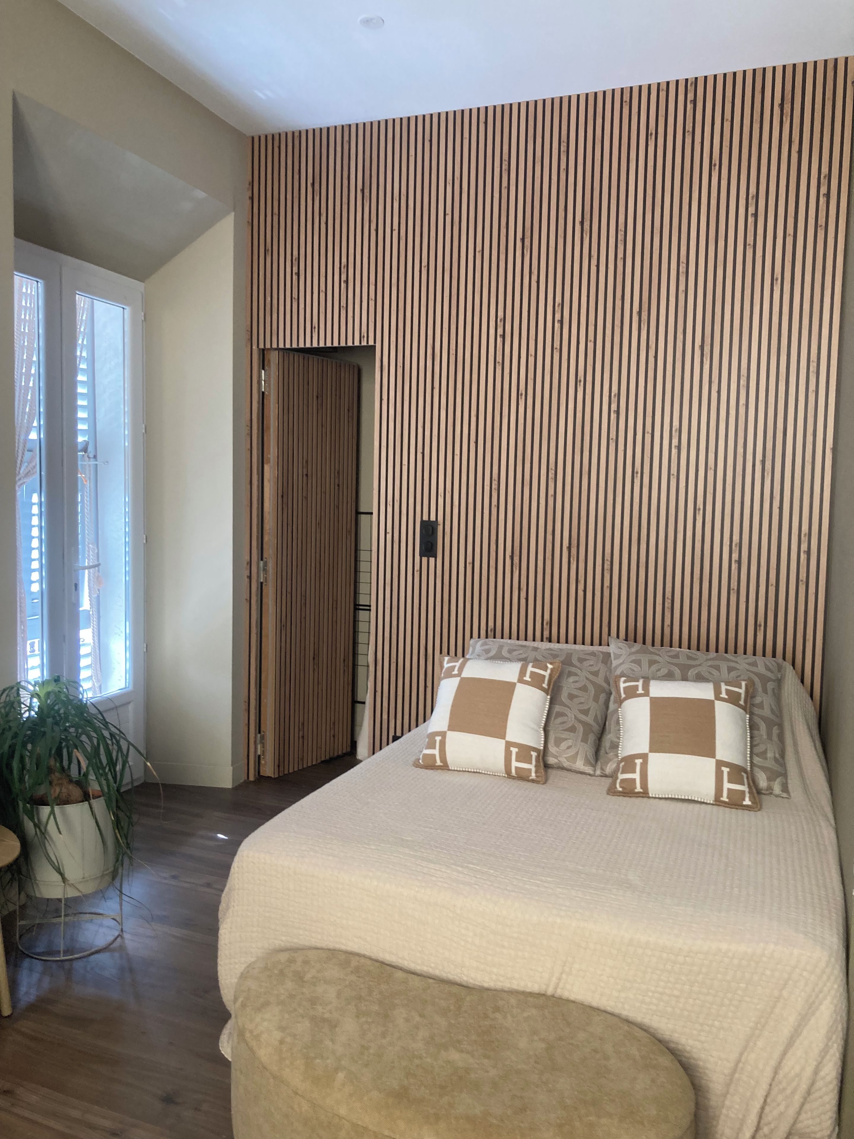 Ein Schlafzimmer mit einem großen Bett, einer Wand aus Holzlatten, einer Topfpflanze und einer Tür zu einem anderen Raum.