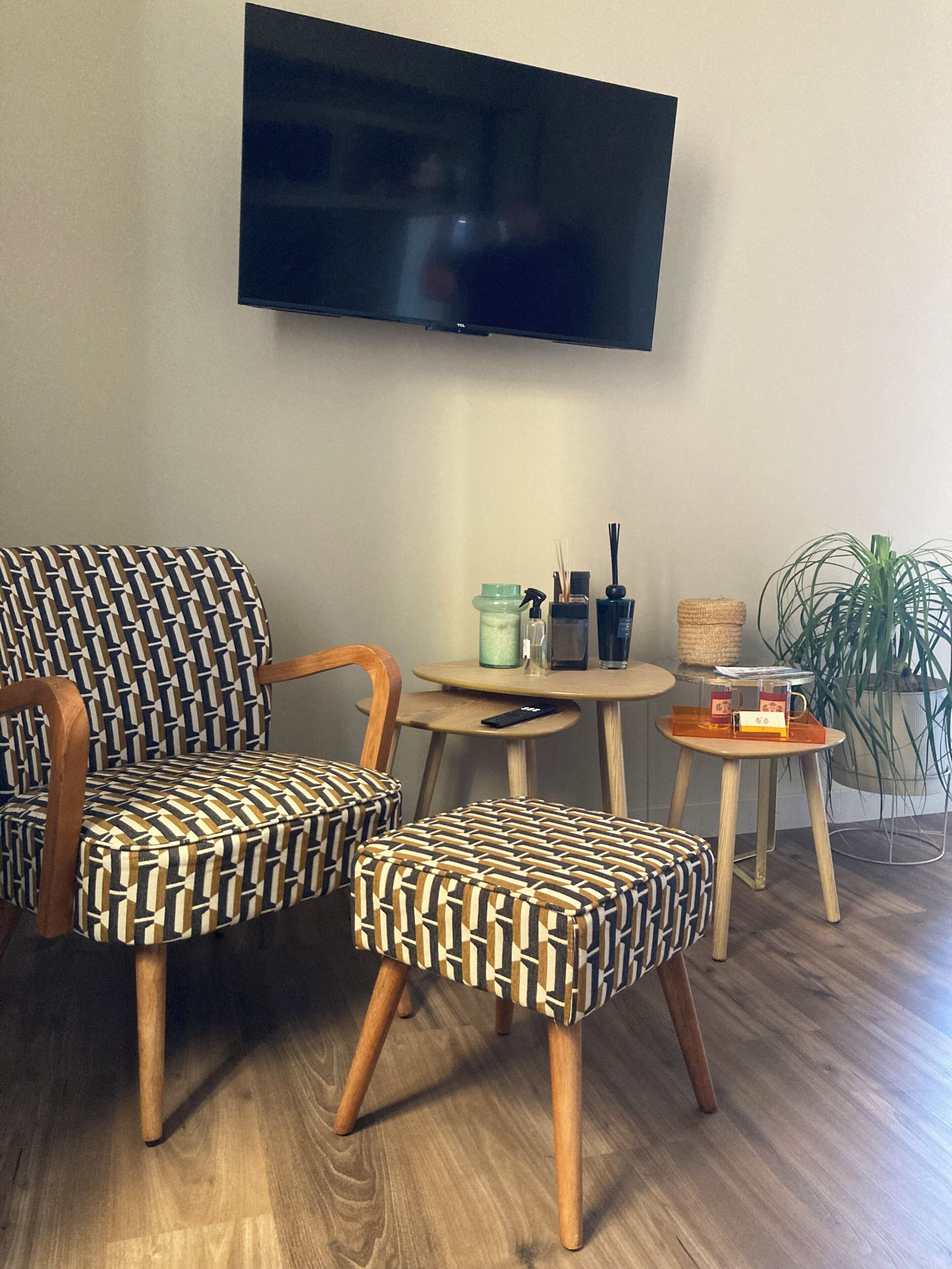 A room with a flat-screen TV mounted on the wall, two patterned armchairs, a small round table with a lamp, a potted plant, and a wooden floor.