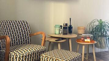 A room with a flat-screen TV mounted on the wall, two patterned armchairs, a small round table with a lamp, a potted plant, and a wooden floor.