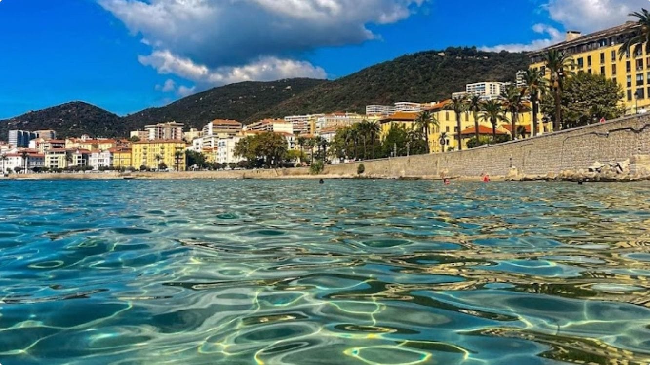 A coastal cityscape with clear waters, buildings, and a hilly backdrop.