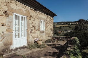 A rustic stone building with two white-framed windows, a small outdoor table, and a potted plant.