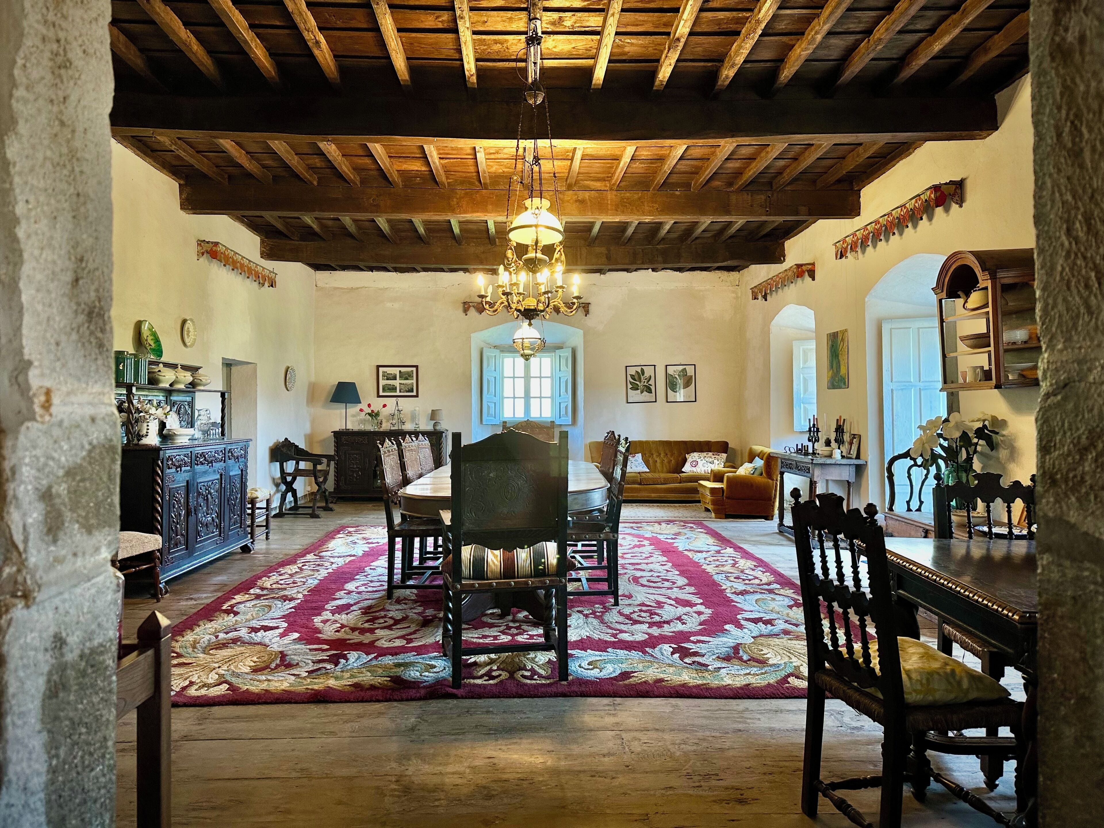Une pièce spacieuse avec un plafond en bois, un lustre, une table à manger avec des chaises, un canapé et un tapis à motifs.