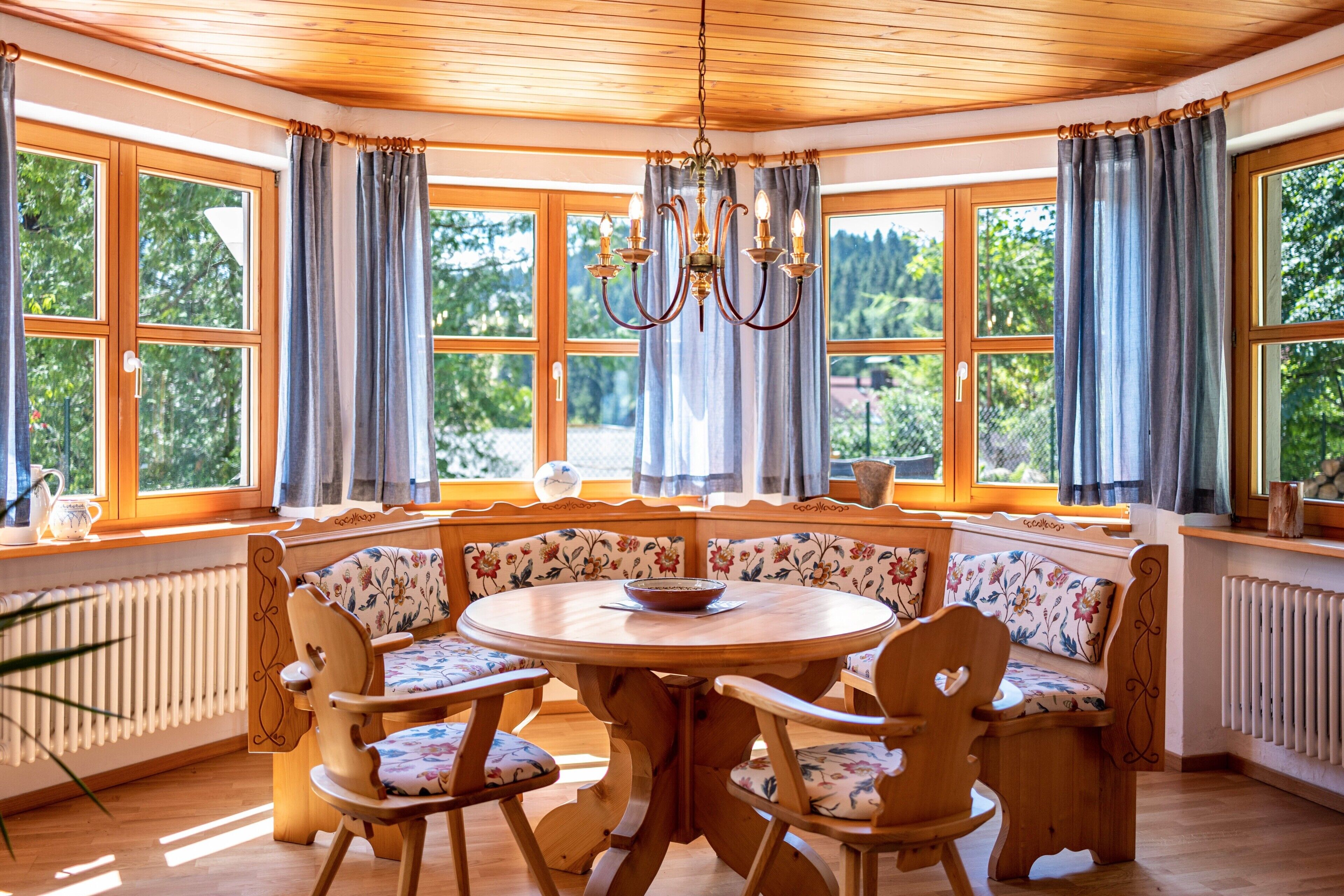 A cozy room with a round wooden table, floral-patterned chairs, and a bench. Large windows with curtains allow natural light and views of greenery.