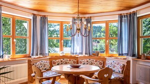 A cozy room with a round wooden table, floral-patterned chairs, and a bench. Large windows with curtains allow natural light and views of greenery. - Apartment "Time Out in the Black Forest" with Private Terrace, Garden & Wi-Fi (Schonach im Schwarzwald)