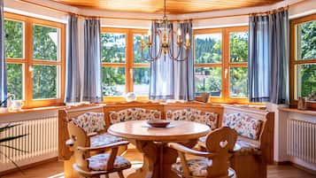 A cozy room with a round wooden table, floral-patterned chairs, and a bench. Large windows with curtains allow natural light and views of greenery.