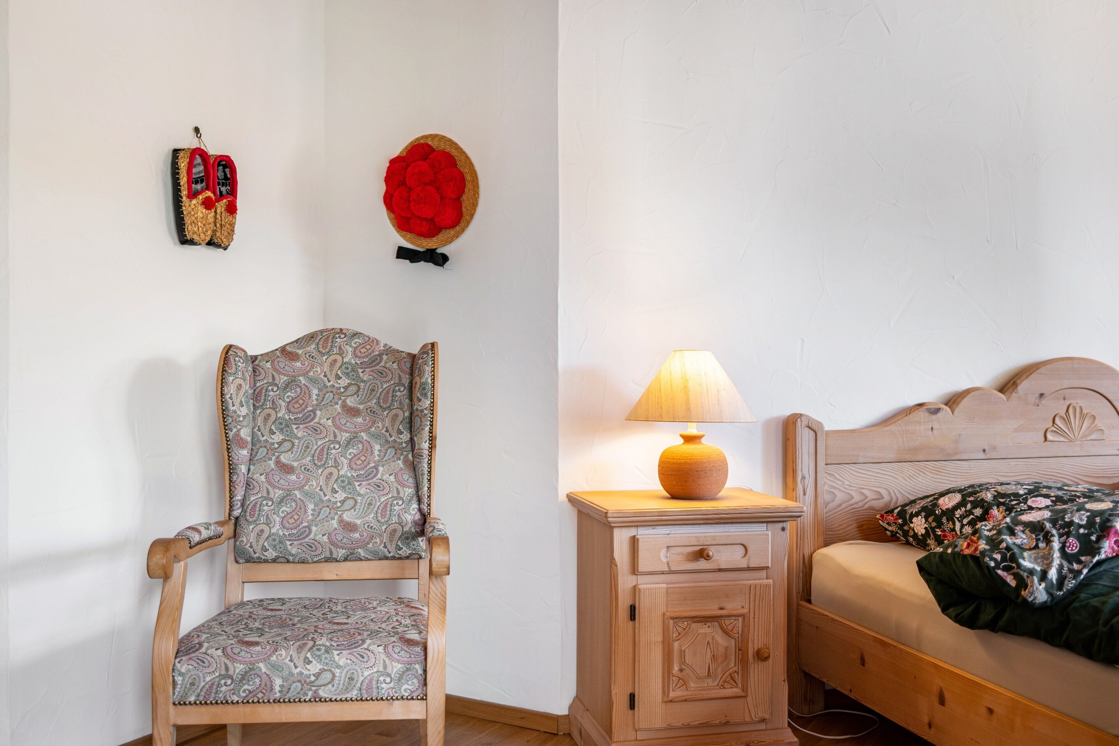 Une pièce comprenant une chaise en bois, une petite table de chevet en bois avec une lampe, et un lit recouvert d’une courtepointe à motifs floraux.