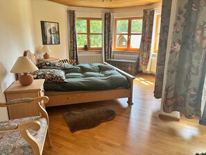 A wooden bed with a green bedspread, a wooden nightstand with a lamp, a chair with a patterned cushion, a window with curtains, and a radiator. - Apartment 'Auszeit Im Schwarzwald' with Private Terrace, Shared Garden and Wi-Fi (Schonach im Schwarzwald)
