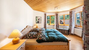 A bedroom with a wooden ceiling, a bed with floral bedding, a nightstand with a lamp, and a window with curtains. - Apartment "Time Out in the Black Forest" with Private Terrace, Garden & Wi-Fi (Schonach im Schwarzwald)