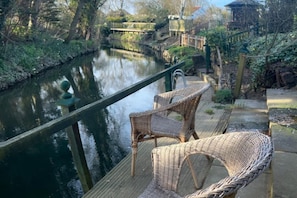 Eine Holzterrasse mit Korbstühlen mit Blick auf einen ruhigen Fluss, umgeben von Grün und einer kleinen Holzhütte.