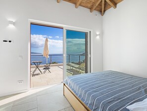 Ein Schlafzimmer mit einem Bett, einer Schiebetür zu einer Terrasse im Freien mit Meerblick und einer Decke mit freiliegenden Holzbalken.
