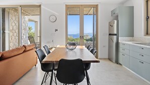 A modern kitchen with a large island, a dining area with a wooden table and chairs, and a view of the sea. - Villa Thymari, new cocoon of privacy and peace (sivota village)