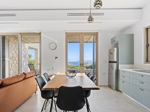 A modern kitchen with a large island, a dining area with a wooden table and chairs, and a view of the sea. - Villa Thymari, new cocoon of privacy and peace (sivota village)