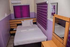 Un lit simple avec une tête de lit, une table de chevet, une petite table, une fenêtre avec des stores violets et un climatiseur fixé au mur.