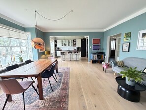 Ein geräumiger Wohnbereich mit einem Holzesstisch, einer Couch und einem Couchtisch. Im Hintergrund ist eine Küche mit weißen Schränken und einer Bar zu sehen.