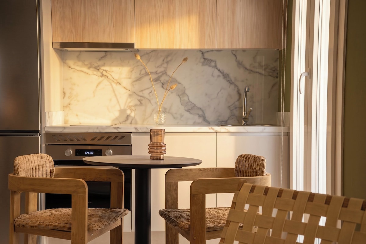 A modern kitchen with a marble backsplash, a built-in oven, and a dining area with two wicker chairs and a round table.