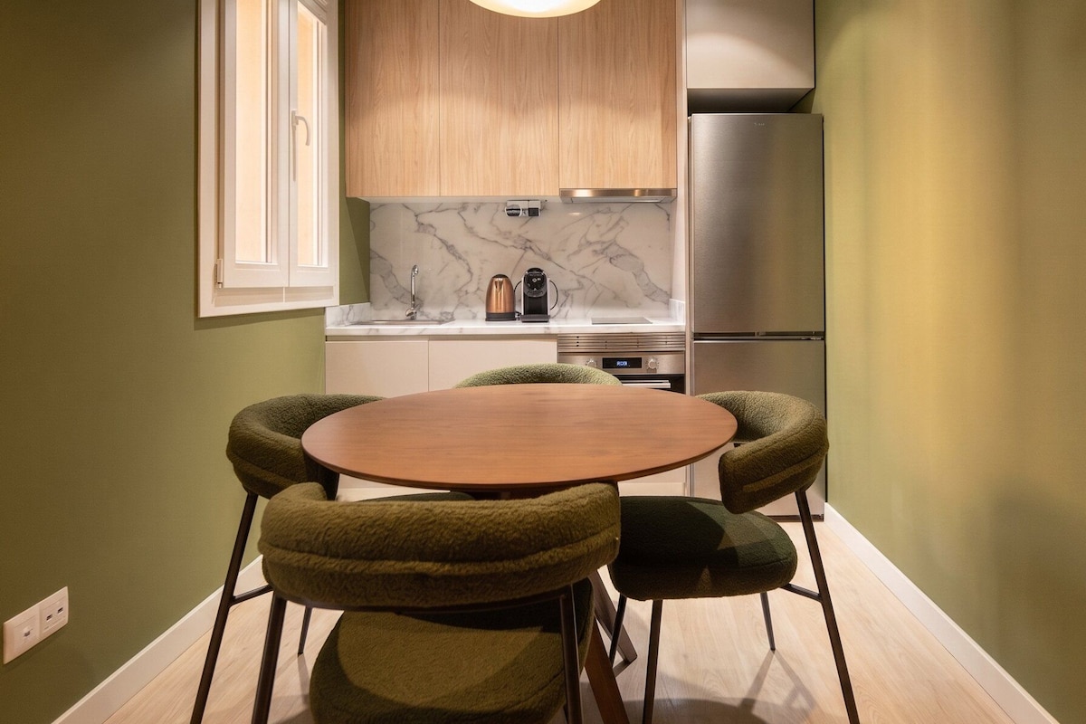 A compact kitchen with a round wooden table and matching chairs, a built-in oven, and a stainless steel refrigerator.