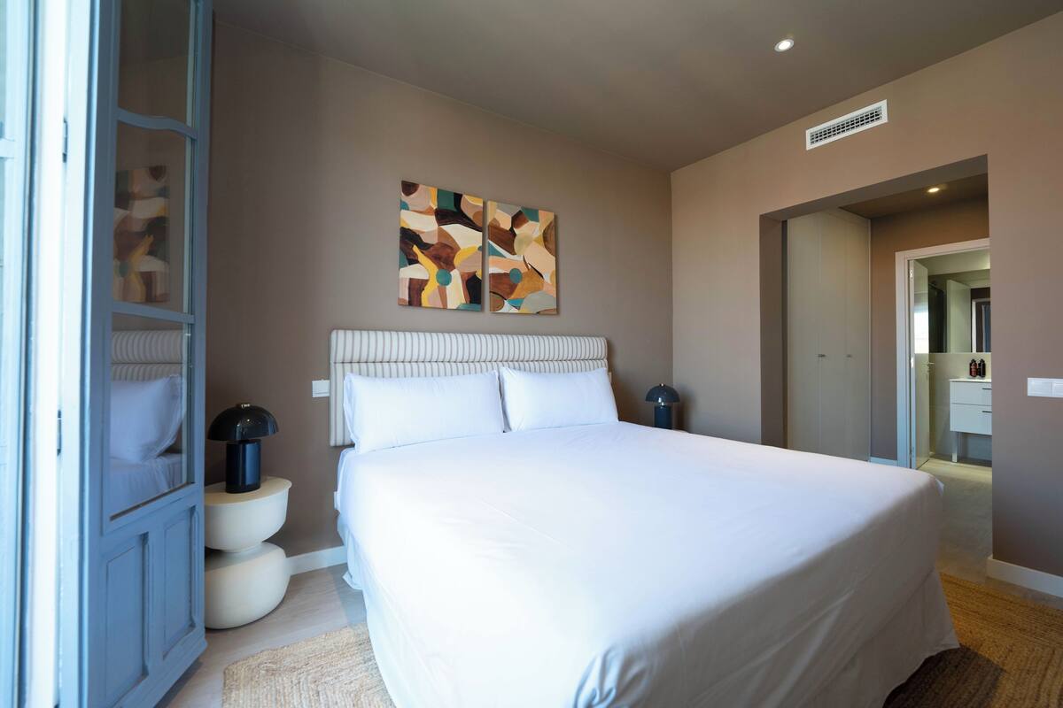 A bedroom with a large bed, a bedside table, a lamp, a wall art piece, and a wardrobe.