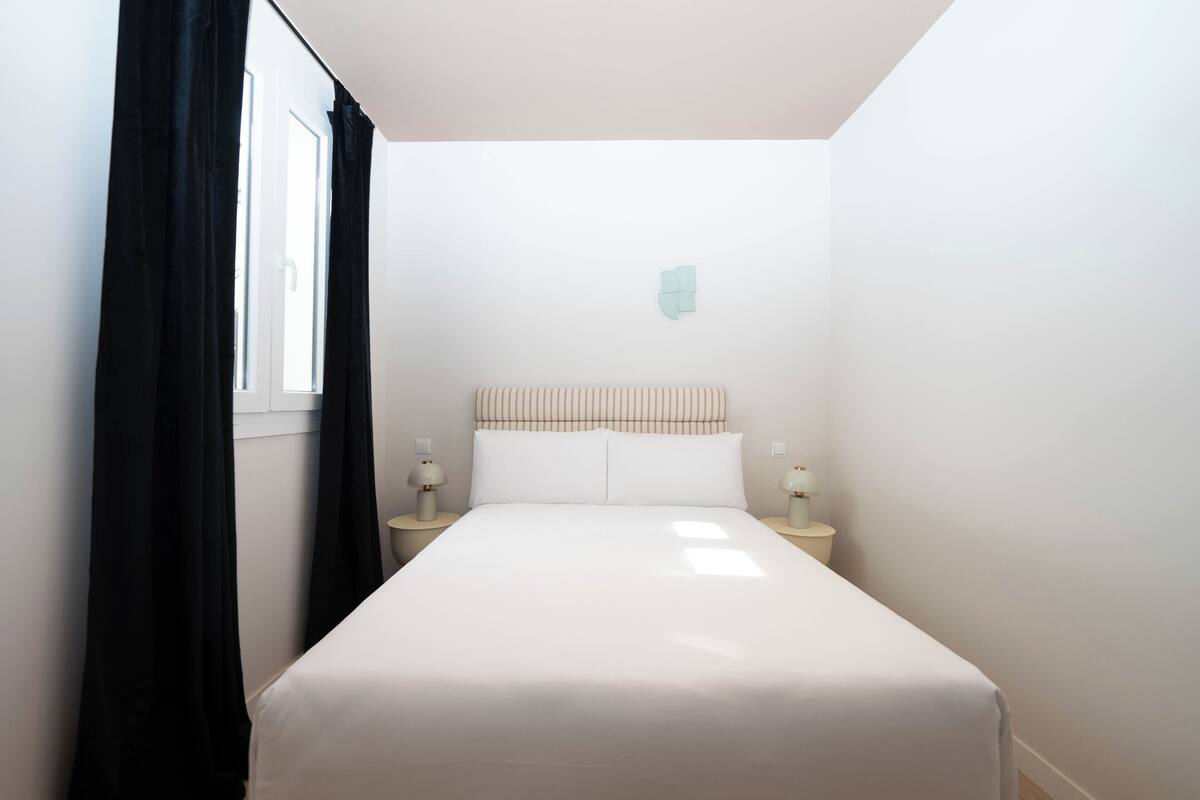 A single bed with a white headboard, two bedside tables with lamps, and a window with black curtains.