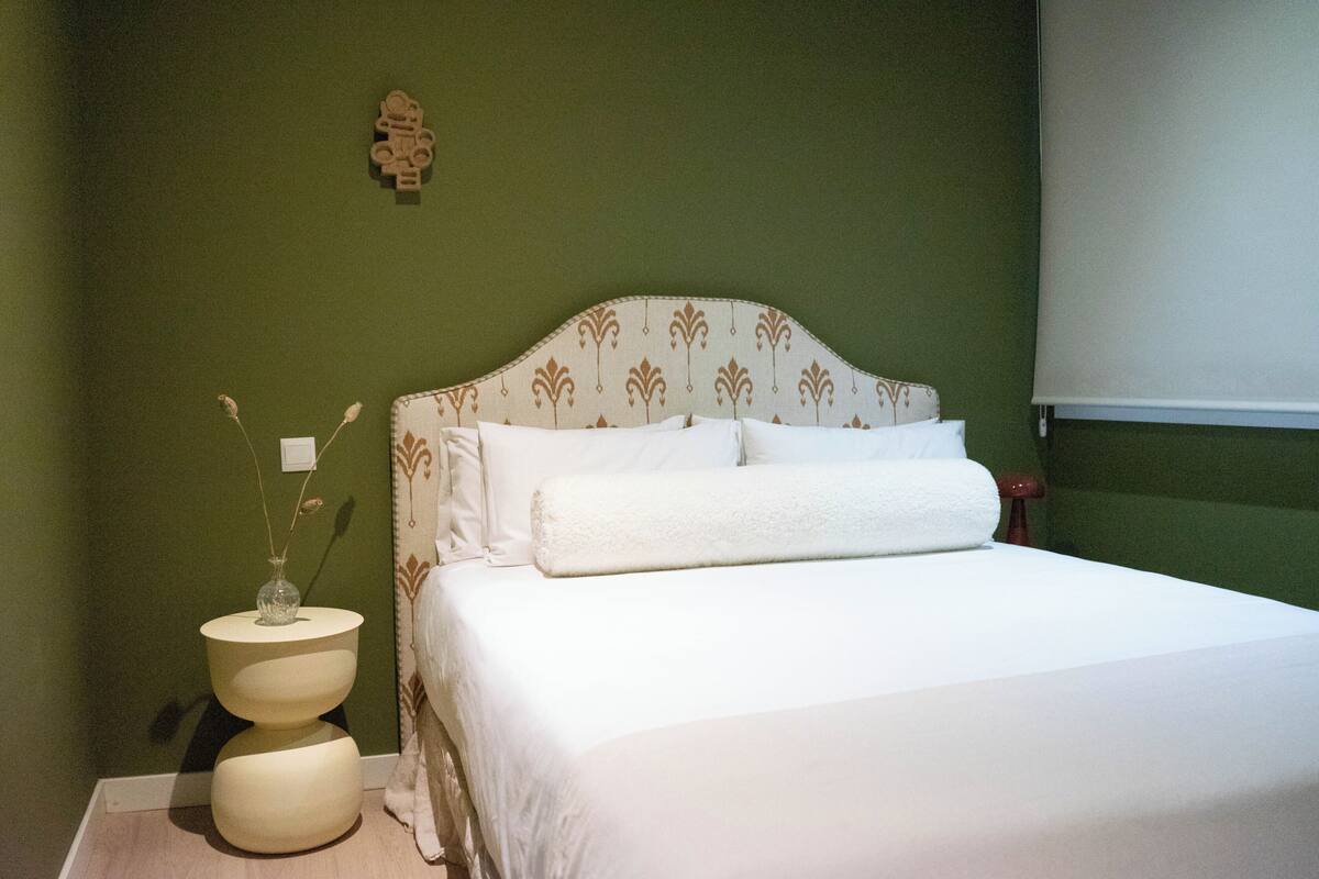 A bed with white bedding, a bedside table with a vase, and a wall-mounted clock.