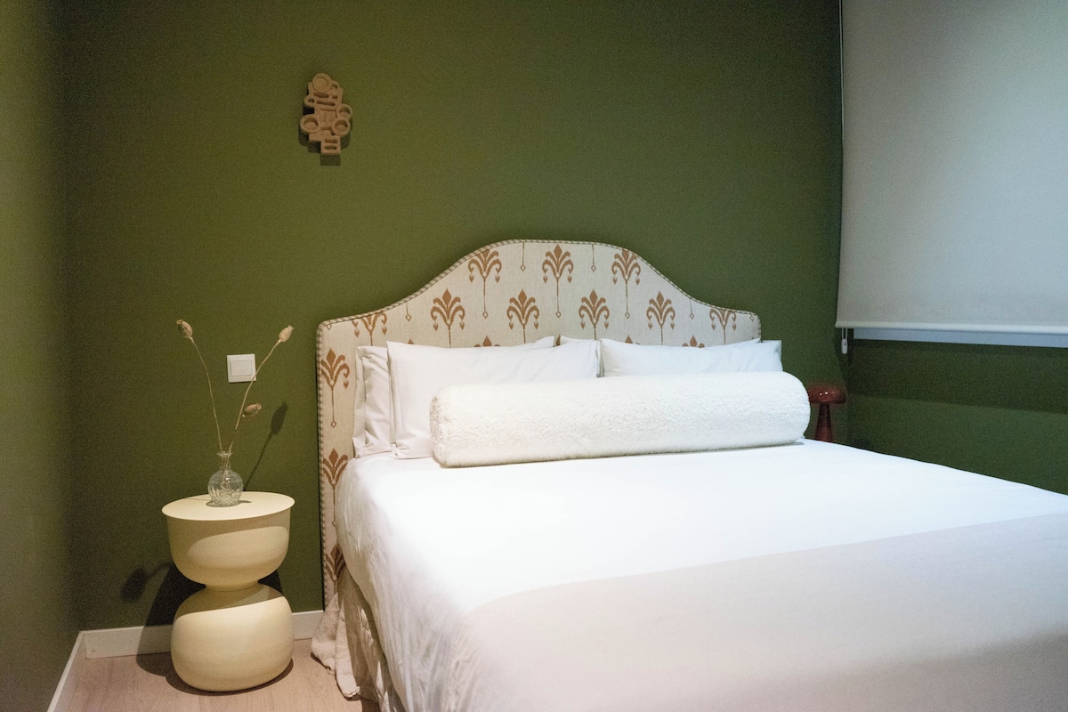 Ein Bett mit weißer Bettwäsche, ein Nachttisch mit einer Vase und eine an der Wand hängende Uhr.