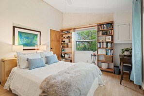 A bedroom with a large bed, a wooden bookshelf, a desk, and a window with a view of trees.