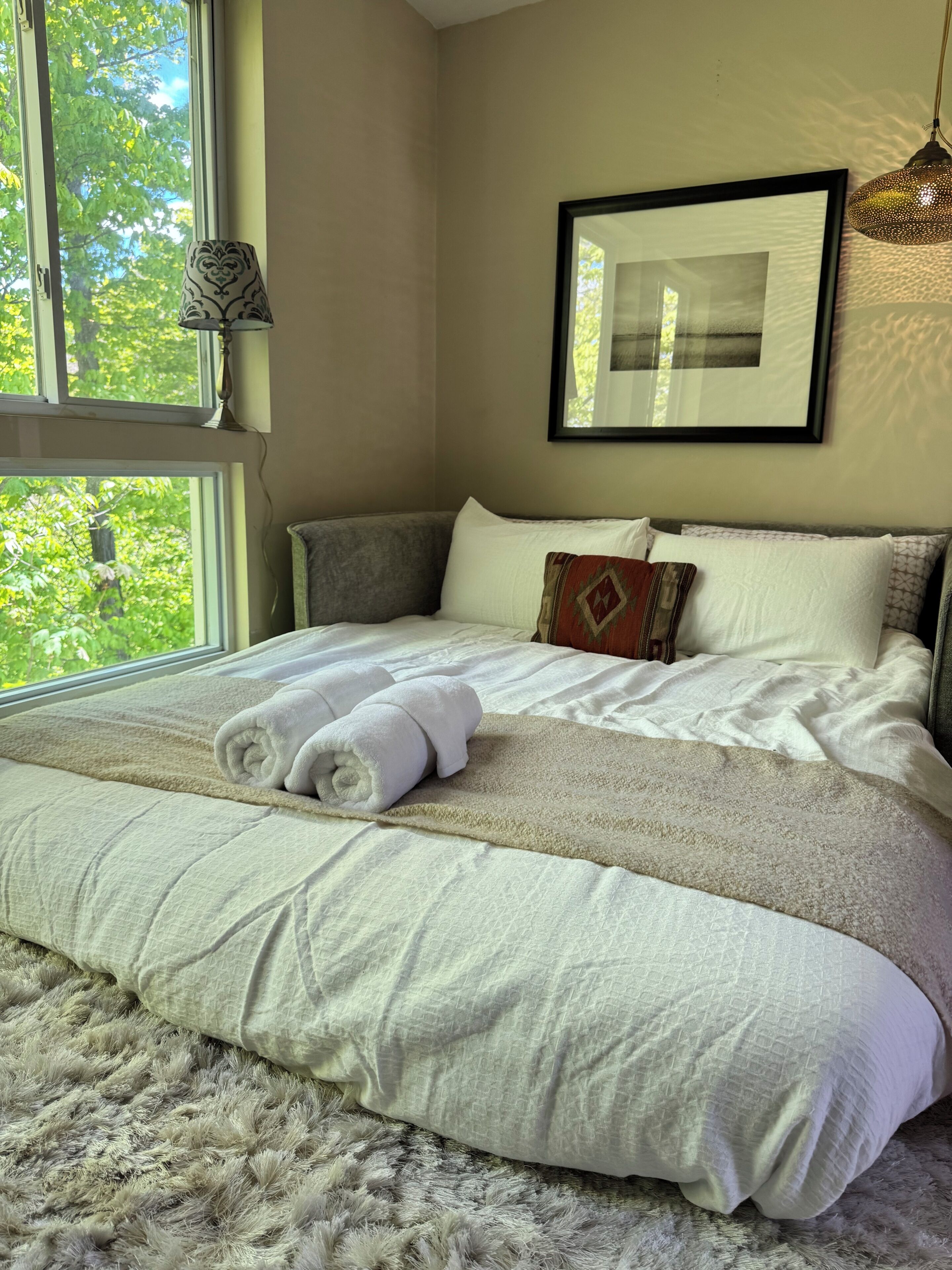 A neatly made bed with a rolled white towel, a framed picture on the wall, and a window with a view of trees.