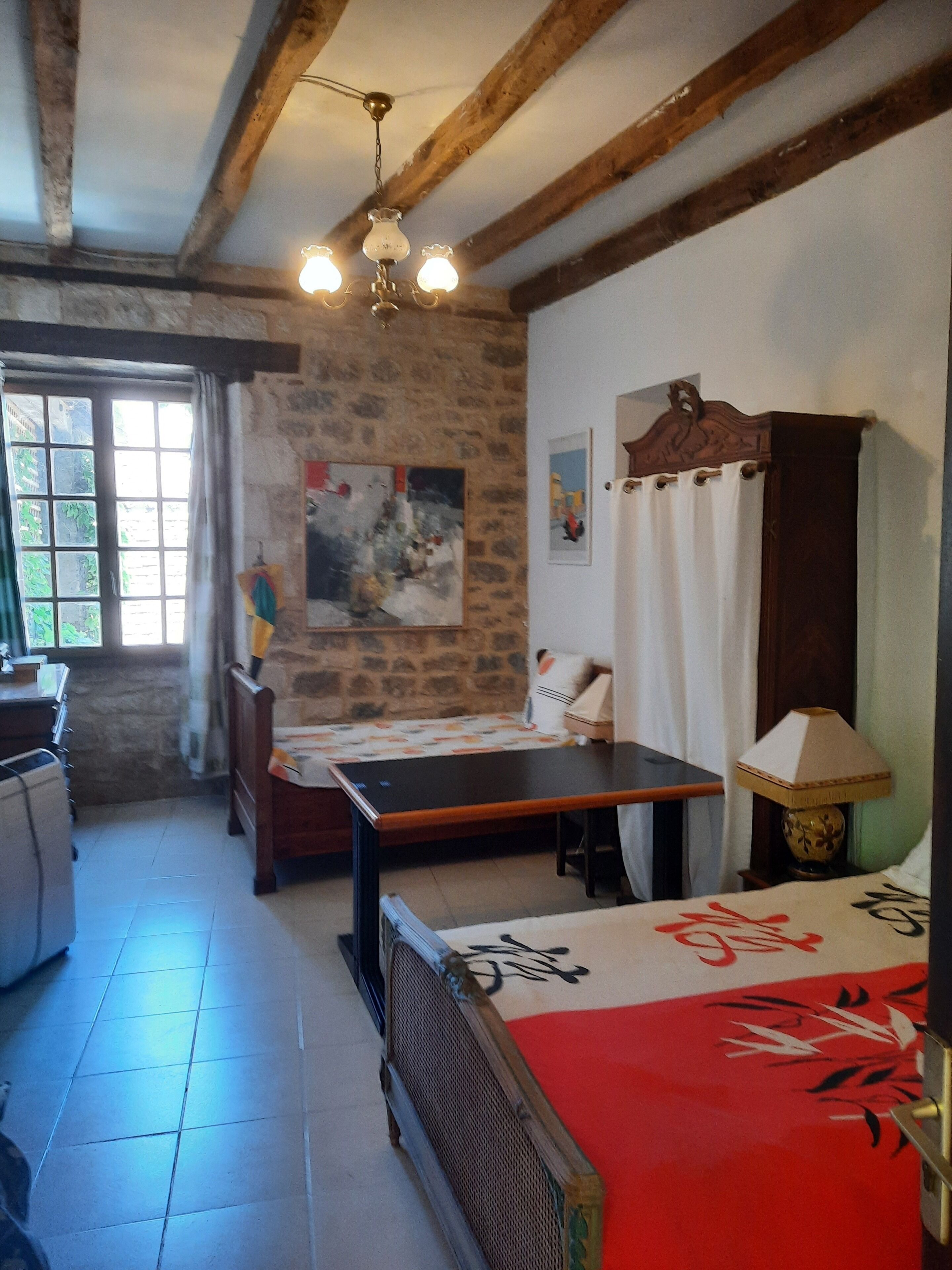Una habitación con cama, una mesa, una silla, una lámpara, una ventana con cortinas, un cuadro en la pared y una pared de piedra.