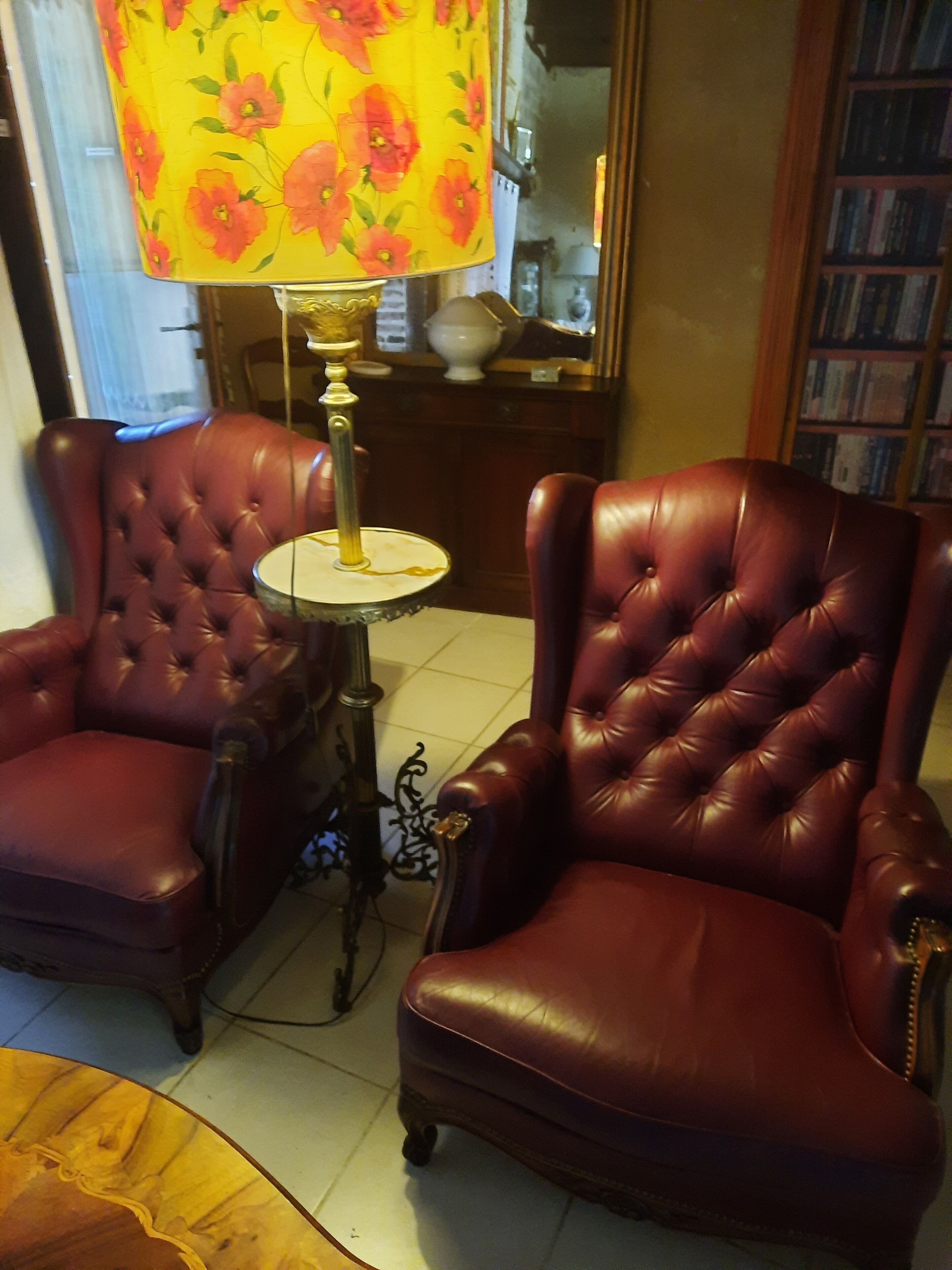 Dos sillones de cuero, una mesita redonda y una pantalla de lámpara con estampado floral en una habitación con suelo de baldosas y una estantería al fondo.