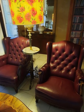 Twee leren fauteuils, een kleine ronde tafel en een bloemenlampenkap in een kamer met een betegelde vloer en een boekenplank op de achtergrond.