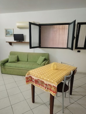 Ein Zimmer mit einem grünen Sofa, einem Esstisch mit gelb-kariertem Tischläufer, einem Fernseher auf einem Regal und einem Fenster mit Gittermuster.