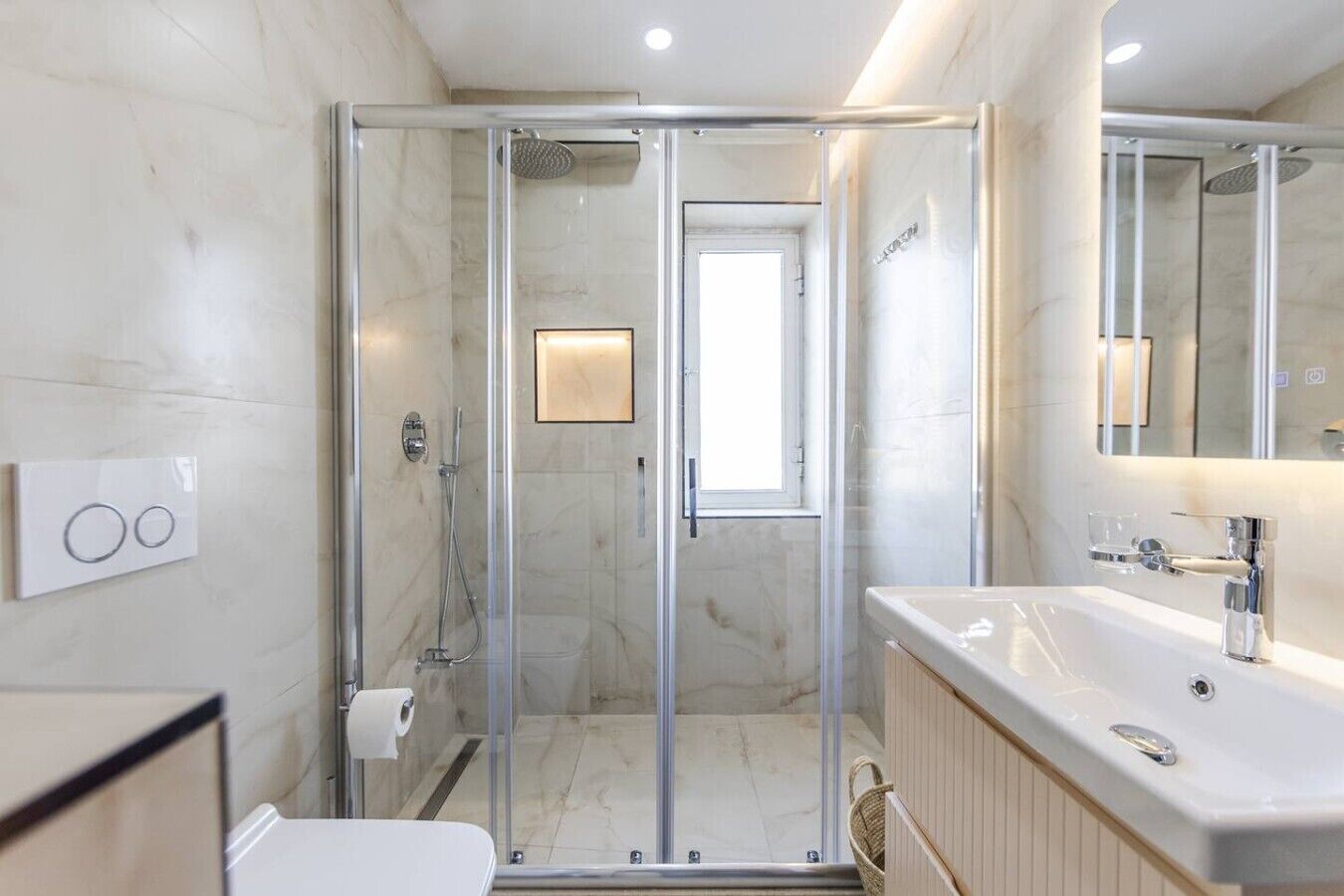 Un baño moderno con ducha envuelta en vidrio, un lavabo blanco con accesorios de cromo y un espejo sobre el lavabo.