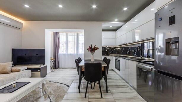 A modern living room with a flat-screen TV, a sofa, a dining table with chairs, and a kitchen with a stainless steel refrigerator. - Shkodra Living | Modern 1BR Near City Center by PikHost (Shkodër)