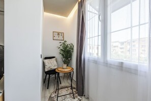 Habitació d'angle amb finestra, una cadira, una taula petita, una planta i un certificat emmarcat a la paret.