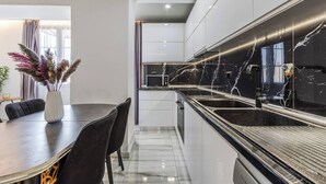 A modern kitchen with a black marble countertop, white cabinetry, and a dining area with a dark table and chairs. - Shkodra Living | Modern 1BR Near City Center by PikHost (Shkodër)