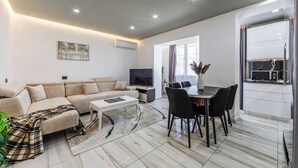 A modern living room with a sofa, coffee table, TV, dining table with chairs, and a kitchen area with a microwave. - Shkodra Living | Modern 1BR Near City Center by PikHost (Shkodër)