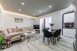 Ein modernes Wohnzimmer mit Sofa, Couchtisch, Fernseher, Esstisch mit Stühlen und einer Küchenzeile mit Mikrowelle.