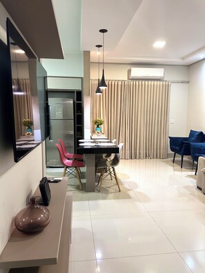 A modern living room with a dining area, a flat-screen TV, a refrigerator, a pendant light, and a large window with curtains. - Davisis Luxo Piscina Prox Graciosa (Palmas)